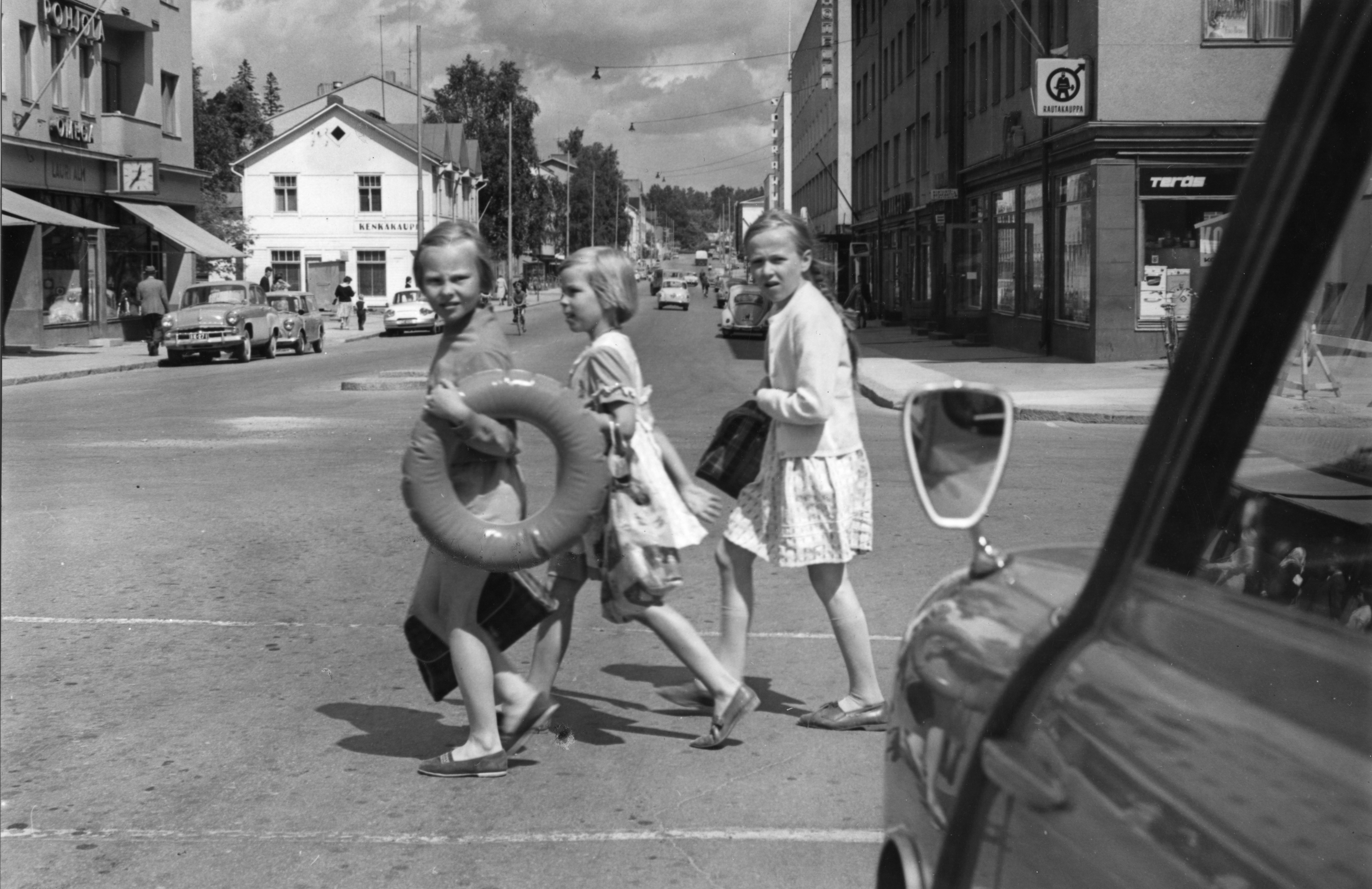 Vanha mustavalkoinen valokuva jossa kolme uimaan menossa olevaa tyttöä ylittää suojatietä uimarenkaat ja pyyhkeet mukanaan.