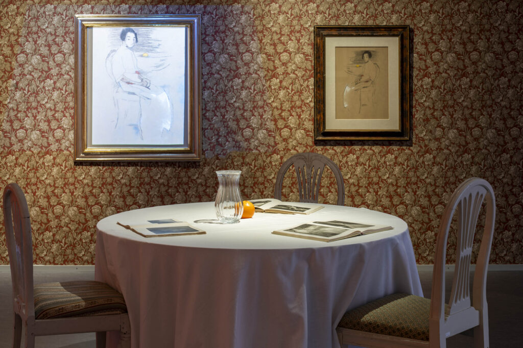 Näyttelysali jossa kukalliset tapetit, maalauksia seinillä sekä pöytä jonka päällä asetelma.